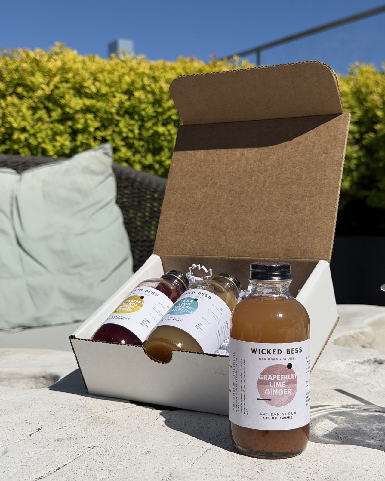 Wicked Bess Mule shrub bundle in an open gift box on a sunny patio, with a 4oz Grapefruit Lime Ginger shrub in front